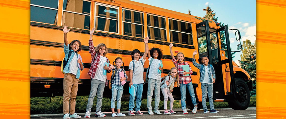 A group of children with backpacks and notebooks wave in front of a yellow school bus