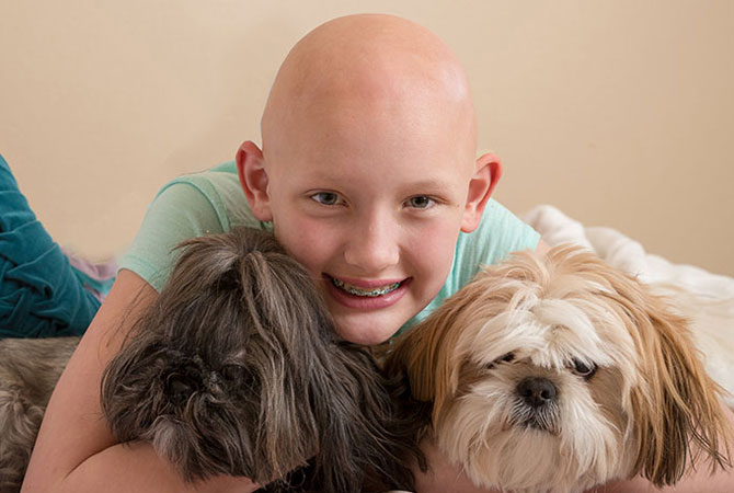 A girl with no hair hugging two dogs